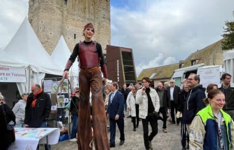 l'animateur micro déambule dans la foire commercial organisée par la communauté de communes de Houdan. Il est entouré d'un décors haut en couleur avec la Tour du Donjon de Houdan. Le président de la communauté de communes est à ses côtés. Une personne sur échasse est également dans la photo et témoigne de l'attractivité de l'événement.