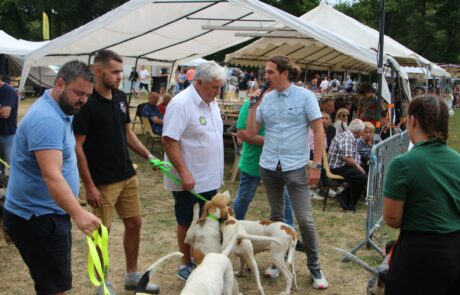 L'animateur tend le micro à une personne durant son interview. Des chiens sont visibles à leurs pieds.