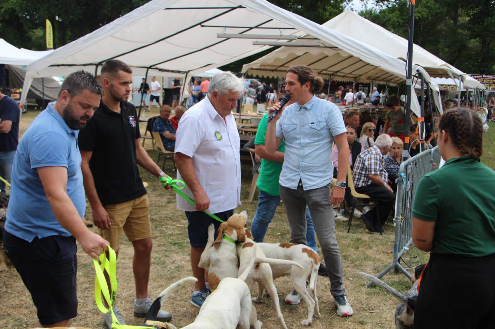 L'animateur tend le micro à une personne durant son interview. Des chiens sont visibles à leurs pieds.